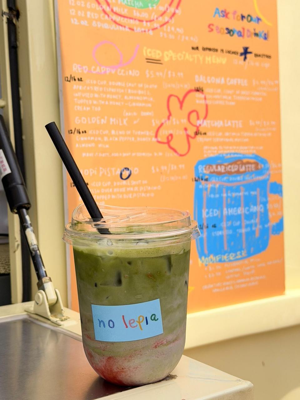 Iced matcha latte with a no lepia sticker in front of the hand-painted menu board